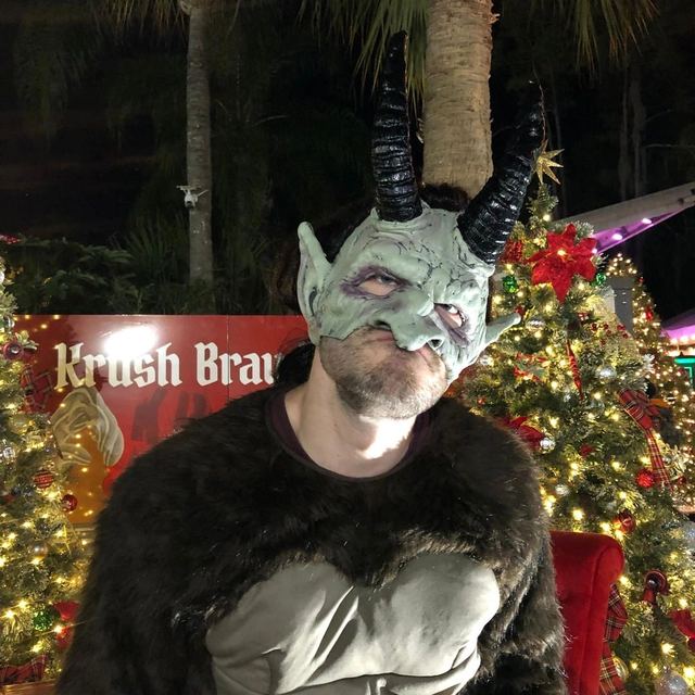Person wearing a horned demon mask and fur cloak in a festive setting.
