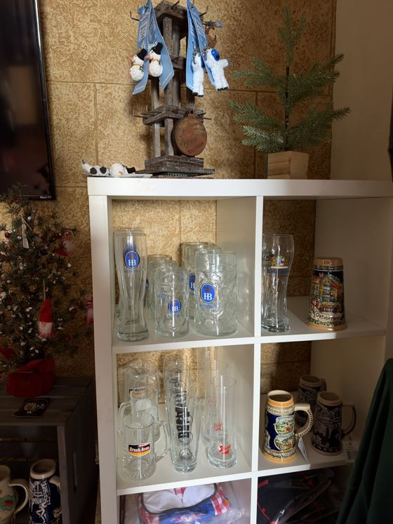 White shelf of Hofbräuhaus HB glass mugs and weizen glasses, a Krush Brau glass mug, a Siegel glass and decorative ceramic steins