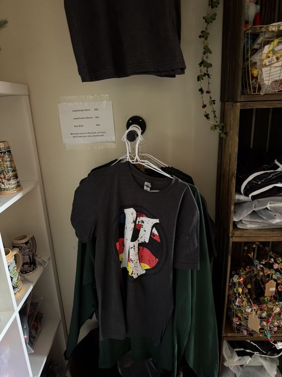 Krush Brau Park apparel wall with a dark gray KB logo tee, steins on a shelf and a sign listing lederhosen shirt $20, shorts $25, both $40