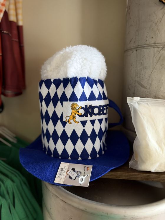 Blue and white Bavarian-diamond felt hat shaped like a beer mug with a white foam top and an Oktoberfest lion patch