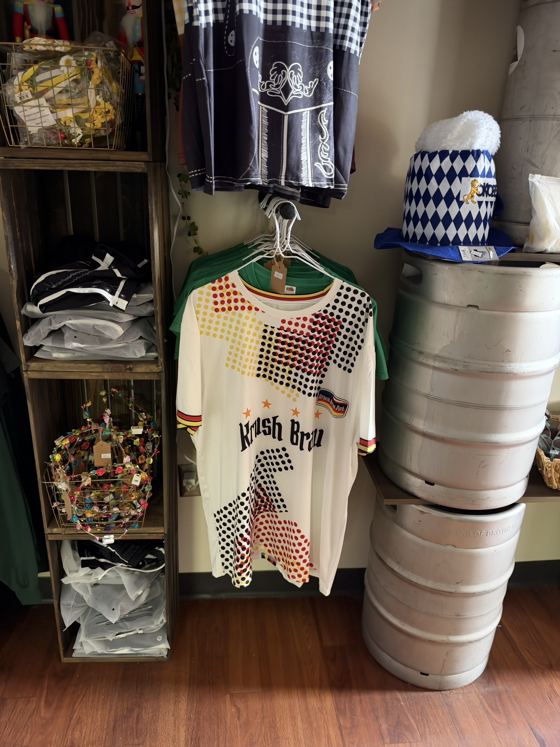 White Krush Brau jersey with black, red and gold German-flag dots, next to flower crowns in a basket and the beer-mug hat on a keg